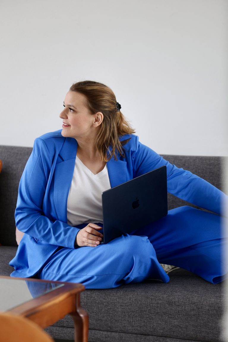 Systemische Beraterin Nora Remstedt sitzt auf dem Sofa und hat ihren Laptop auf dem Schoß sie trägt einenb lauen anzug und schaut nach rechts.