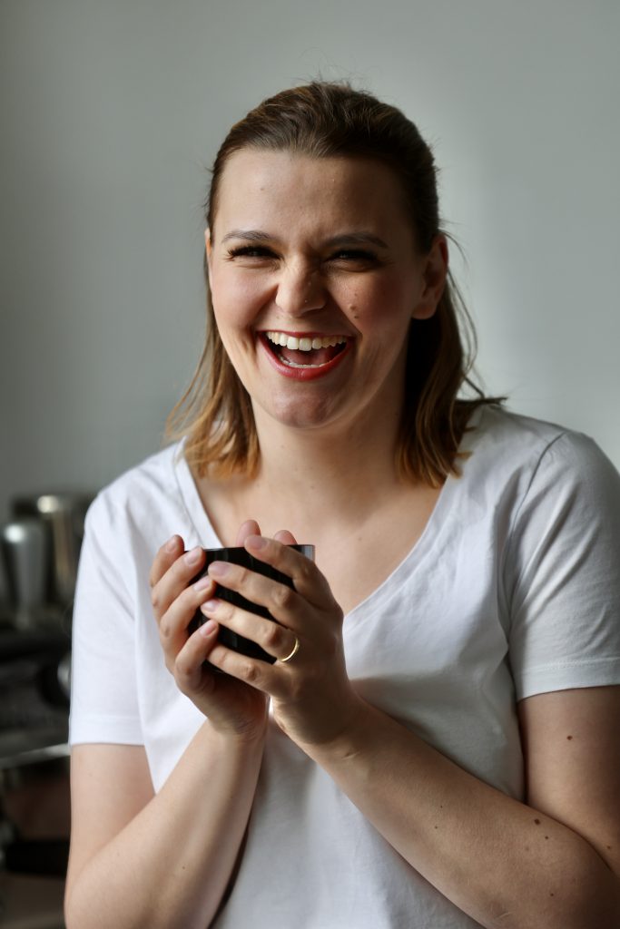 Nora Remstedt persönliche Beratung auf Augenhöhe, steht mit einer Tasse in der Hand und lacht in die Kamera sie trägt ein weißes shirt.
