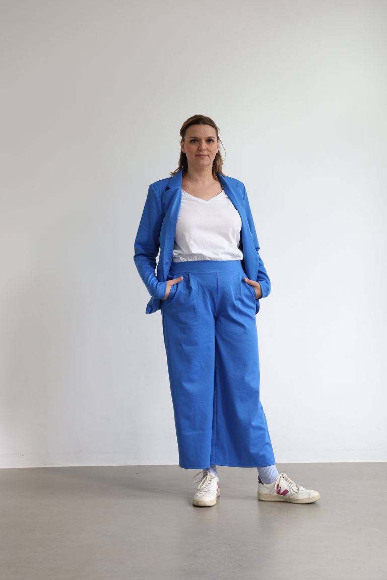 Nora Remstedt referntin für Stressmanagement sthet vor einer weißen Wand im Blauen Anzug und lächelt leicht in die Kamera, ihre Hände sind locker in die Hosentasche gesteckt. Die Haare sind halbofffen und sie trägt weiße sneackers