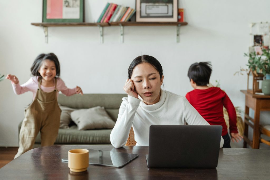 Eine Frau (vermutlich die Mutter) sitzt am Tisch mit einem Laptop und Kafffe wirkt erschöpft und müde zwei Kunder laufen und toben im Hintergund umher. Familien udn Vereinbarkeit von Beruf