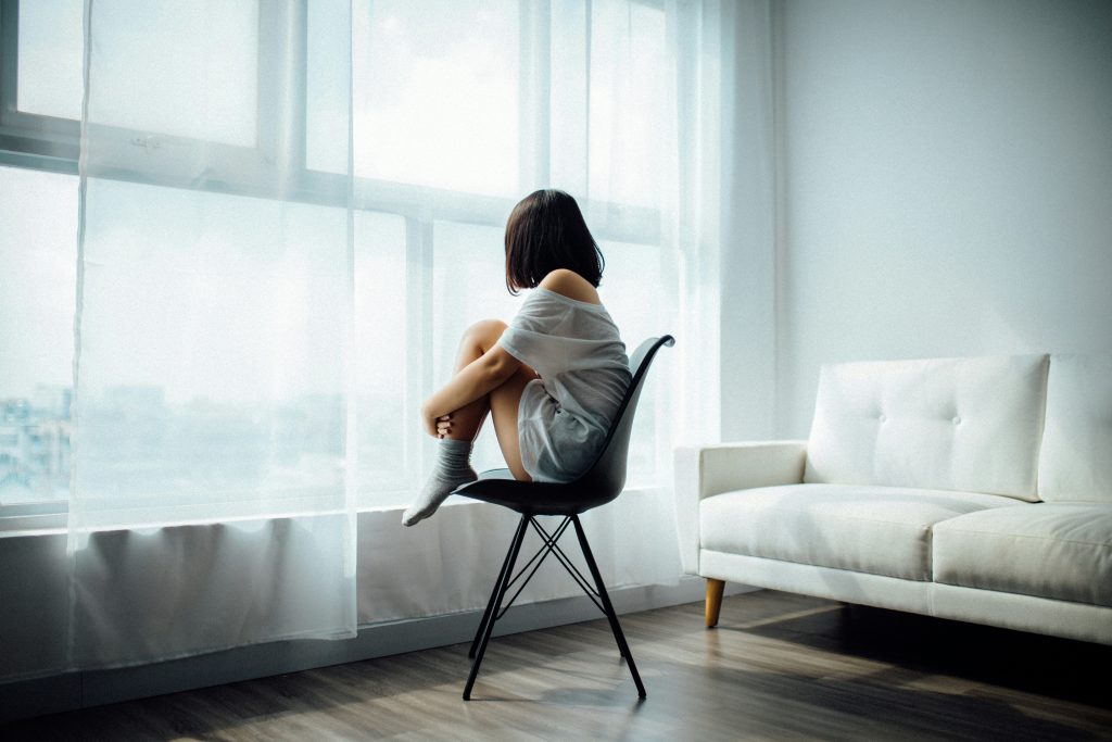 Frau sitzt wegen emotionale erschöpfung auf einem Stuhl Beine angezogen, man erkennt nur die schwarzen Kinnlagen Haare Sie schau durch dünne gardinen nach draußen.